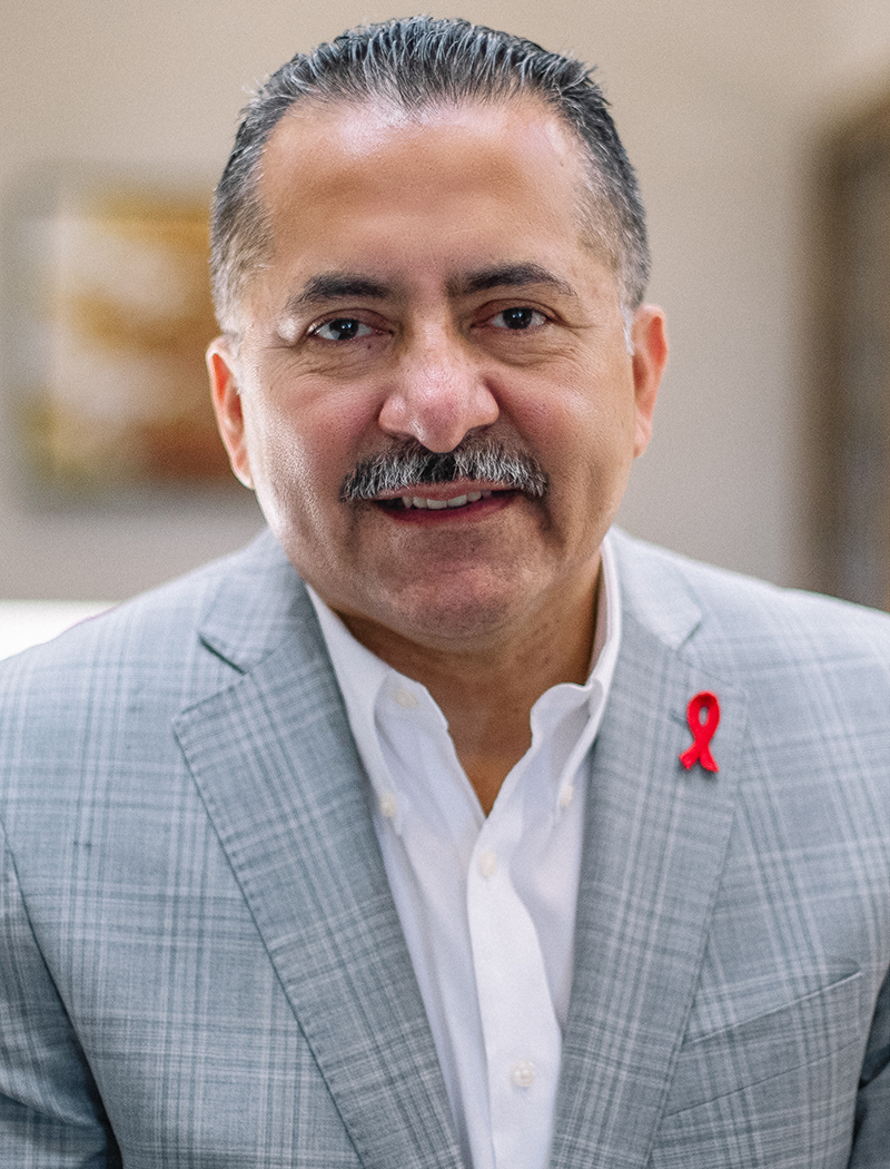 Photo of Guillermo Chacon, latino man with salt and pepper hair, a mustach, wearing a white button down shirt with a plaid jacket; the jacket has a small red AIDS ribbon on the lapel
