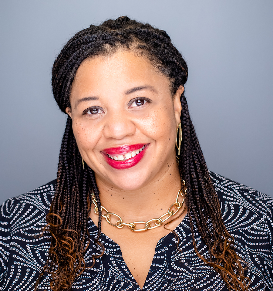 Smiling Black woman in her 30s or 40s, with red lipstick, braids and wearing a pretty chunky gold chain necklace and a black and white patterned dress