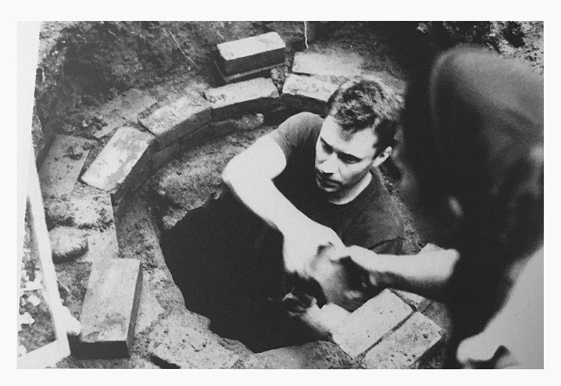 B&W photo of a white man (artist David Nelson) in an old cistern; he's handing something up to someone who's face isn't shown