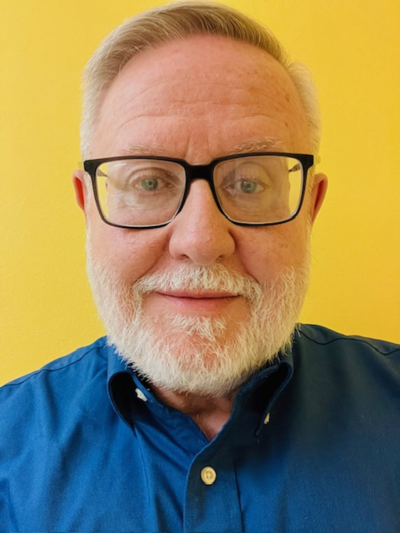 older white man with glasses and a white beard; blue button down shirt against a yellow background